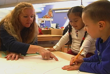 A teacher points to a pathway drawn on paper while two preschoolers observe