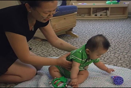 An infant sits on the floor and reaches for a bell while his mother supports him