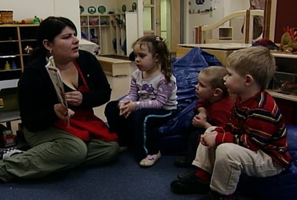 A teacher sits on the floor and reads a picture book to three preschoolers