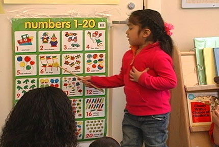 A Latina preschooler uses a pointer to point to numerals on a chart for her classmates