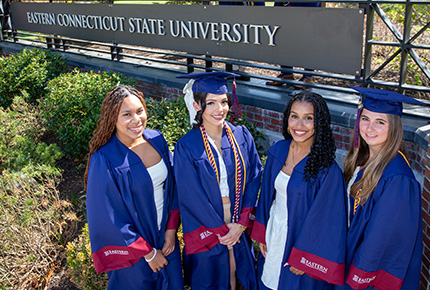 students in cap and gown in front of the Eastern Connecticut State University entrance sign