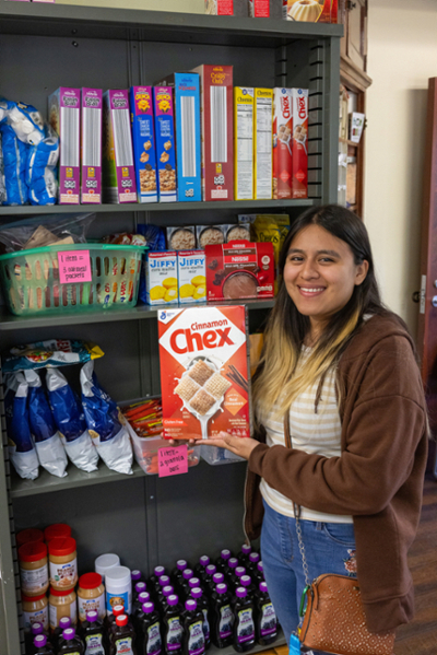 A student standing beside a cupboard loaded with canned goods and other foodstuff