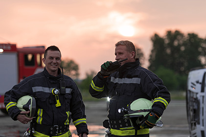 two smiling fireman, one talking on a walkie-talkie with a firetruck in the background