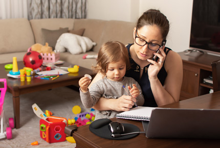 Parent with toddler on lap while talking on phone and looking at laptop. Many toys in background