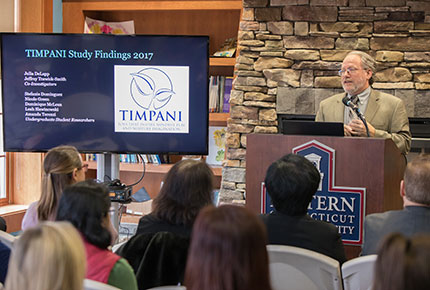 Dr. Trawick-Smith addresses an audience from a podium and gestures toward a large screen with the intro slide about the 2017 TIMPANI study findings