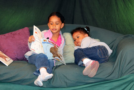 A girl sits on a couch and reads a Spanish picture book to her younger sister