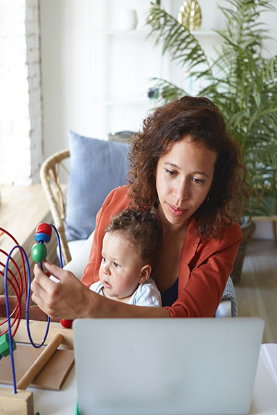 A mother works on a laptop computer with a baby on her lap playing with a toy.