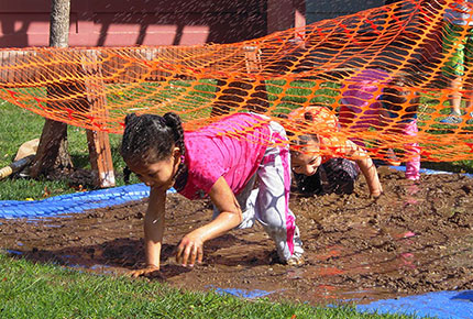 Three preschoolers crawl through the mud under a net