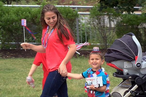 A preschooler and his teacher wear red, white, and blue while marching. The teacher carries a small American flag. A stroller is also visible.