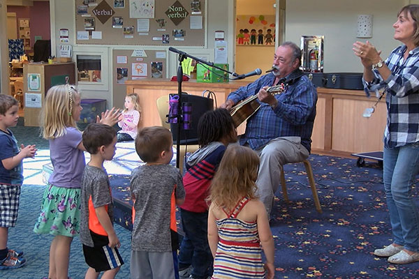 A man sits on a chair on stage, playing a guitar and singing into a microphone. A teacher stands near him, clapping and dancing. Children stand in front of the stage and clap and dance.