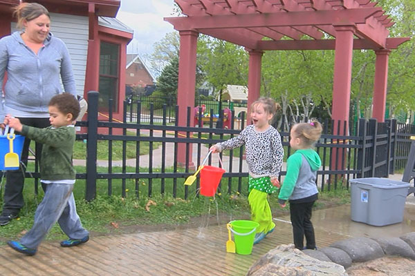 Two toddlers carrying buckets with holes in them, dripping water. Their teacher and a third child observe.