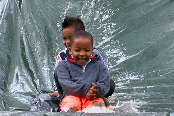 Two smiling preschoolers slide down a hill covered with a tarp and running water