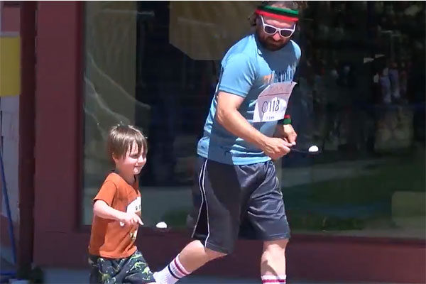 A preschooler and his teacher run, each carrying a plastic egg on a spoon. Both wear numbers attached to their shirts.