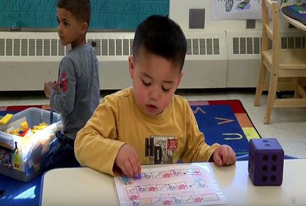 A preschooler advances pieces on a homemade board game. A large die is on the table next to the child.