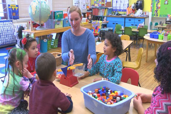 Children and teacher sit around a table, using a balance scale. 