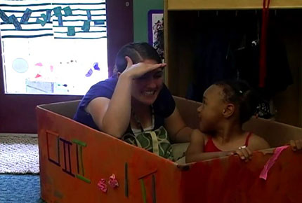 A teacher and student sit inside a box, pretending it's a train.