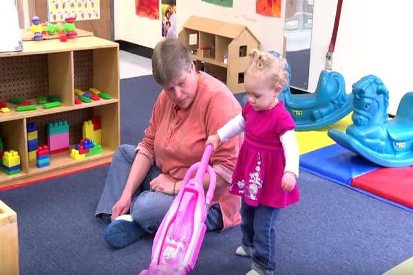 A toddler pushes a toy vacuum as her teacher sits next to her. 