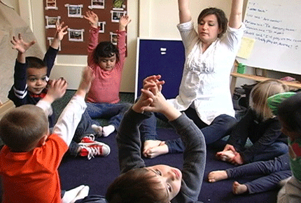 Preschoolers sit on a rug and reach their arms overhead, mimicking their teacher