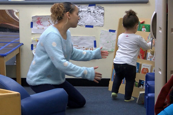 A teacher helps a child pick a book from a bookshelf. 