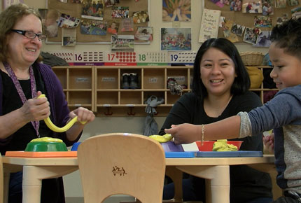 A teacher sits at a table and plays with Gak with a preschooler, while his mother looks on. All are smiling.