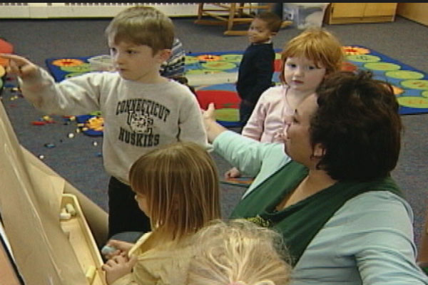 A teacher sits on the floor playing at an easel with young children.