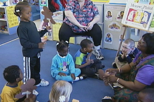 A teacher and children sit in whole group while one child talks to the teacher.
