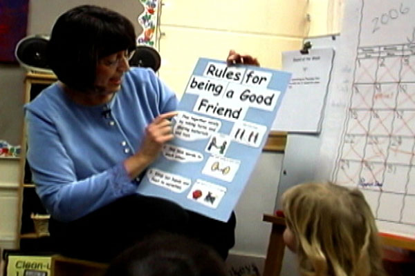 A teacher sits in front of the class and is holding a list of Rules for them to see.