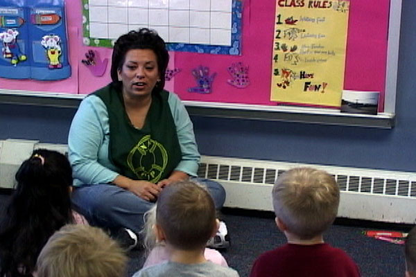 A teacher sits on the floor with a group of children, reviewing classroom rules.