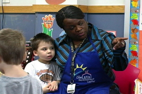 A teacher sits on the floor with a child and points to something.