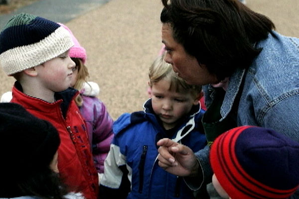 A teacher speaks with a child outside while other children are gathered around.