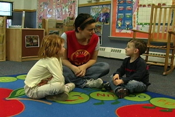 A teacher and two children sit on the floor and talk through a conflict.