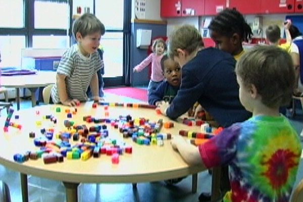 Three children are using blocks on a table and a teacher is helping and speaking to one child.
