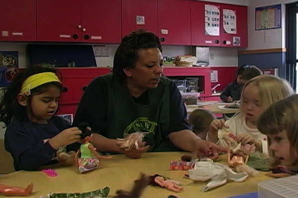 A teacher sits at a table with three children who are playing and she is talking to them.