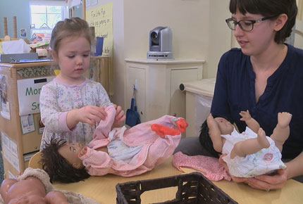 A child dresses a doll while the teacher observes