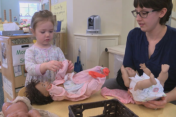 A child dresses a baby doll on a table while her teacher watches.