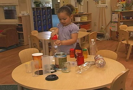 A preschooler stands at a table with a variety of plastic and cardboard bottles and other containers