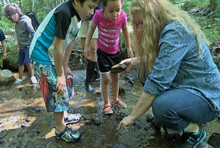 Children gather around their teacher as she lifts over a rock in the forest. 