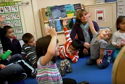 Children sit in a whole group setting looking at a book with their teacher. 