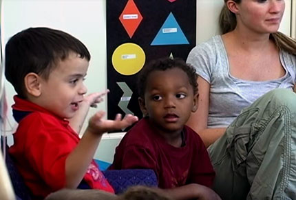 A child sits with another child and an adult on the floor and he is speaking. 