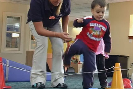 A preschool child steps over a rope strung between two cones as his teacher stands by, ready to assist