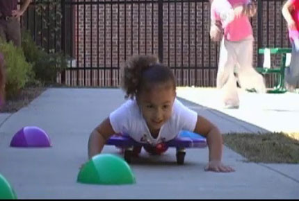 A preschooler lies on her stomach on a scooter and uses her hands to pull herself around a cone on a paved pathway