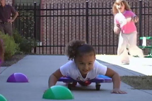 A child lays on her stomach on a scooter and moves through an obstacle course.
