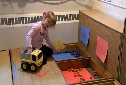 A preschooler drives a dump truck toward bins sorted by color 