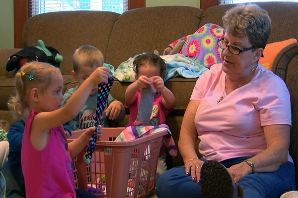 A teacher sits on the floor with toddlers as they sort socks from a laundry basket