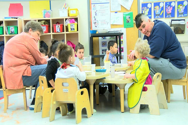 A group of toddlers sit at a table eating lunch with their teachers.