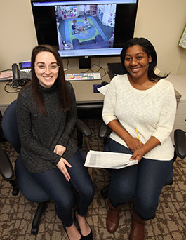 Two student researchers sit in front of a large screen used for research while holding coding sheets