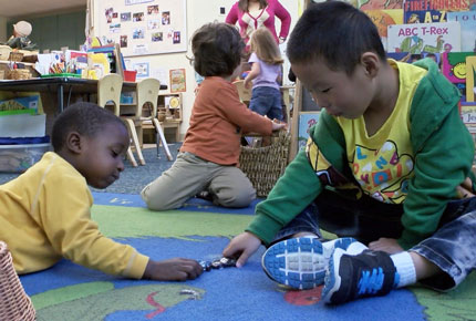 Two preschoolers play with small metal cars on a rug in a classroom