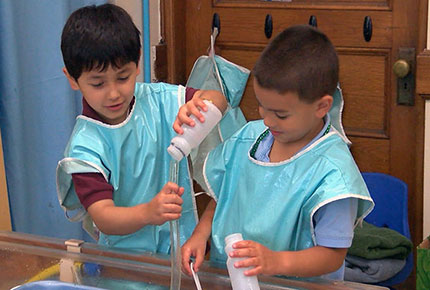 Two preschoolers experiment by pouring water from containers into clear plastic tubing at the water table