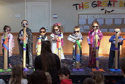Several preschoolers stand on stage in costume, holding hand-made instruments and cardboard microphones