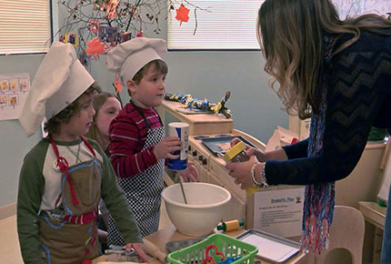 A teacher assists two preschools, dressed in chef hats and aprons, in a play bakery